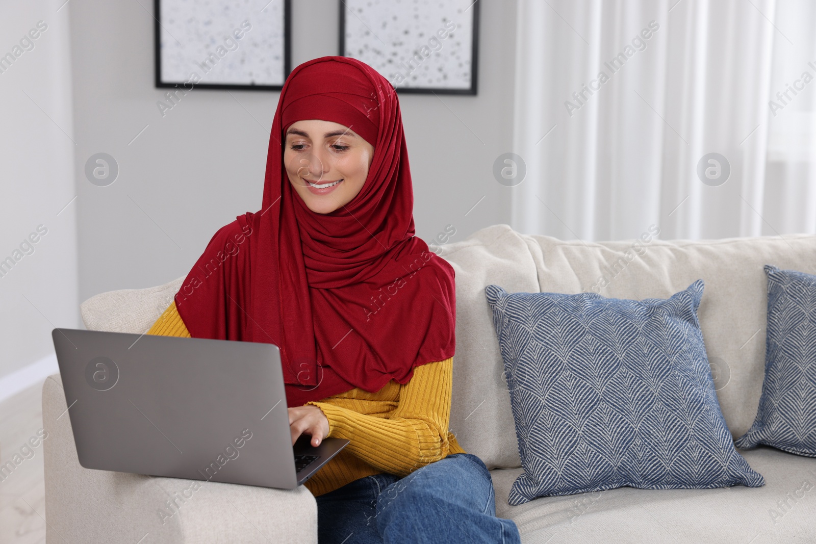 Photo of Muslim woman using laptop at couch in room. Space for text