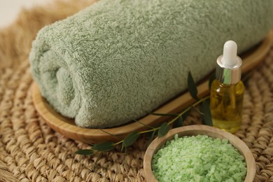 Photo of Spa composition. Rolled towel, cosmetic product, sea salt and twig on wicker mat, closeup