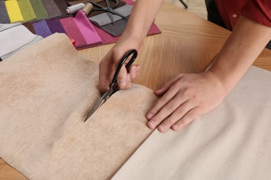 Photo of Man cutting beige fabric with scissors at wooden table, closeup
