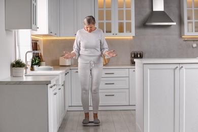 Photo of Menopause, weight gain. Concerned woman standing on floor scales in kitchen