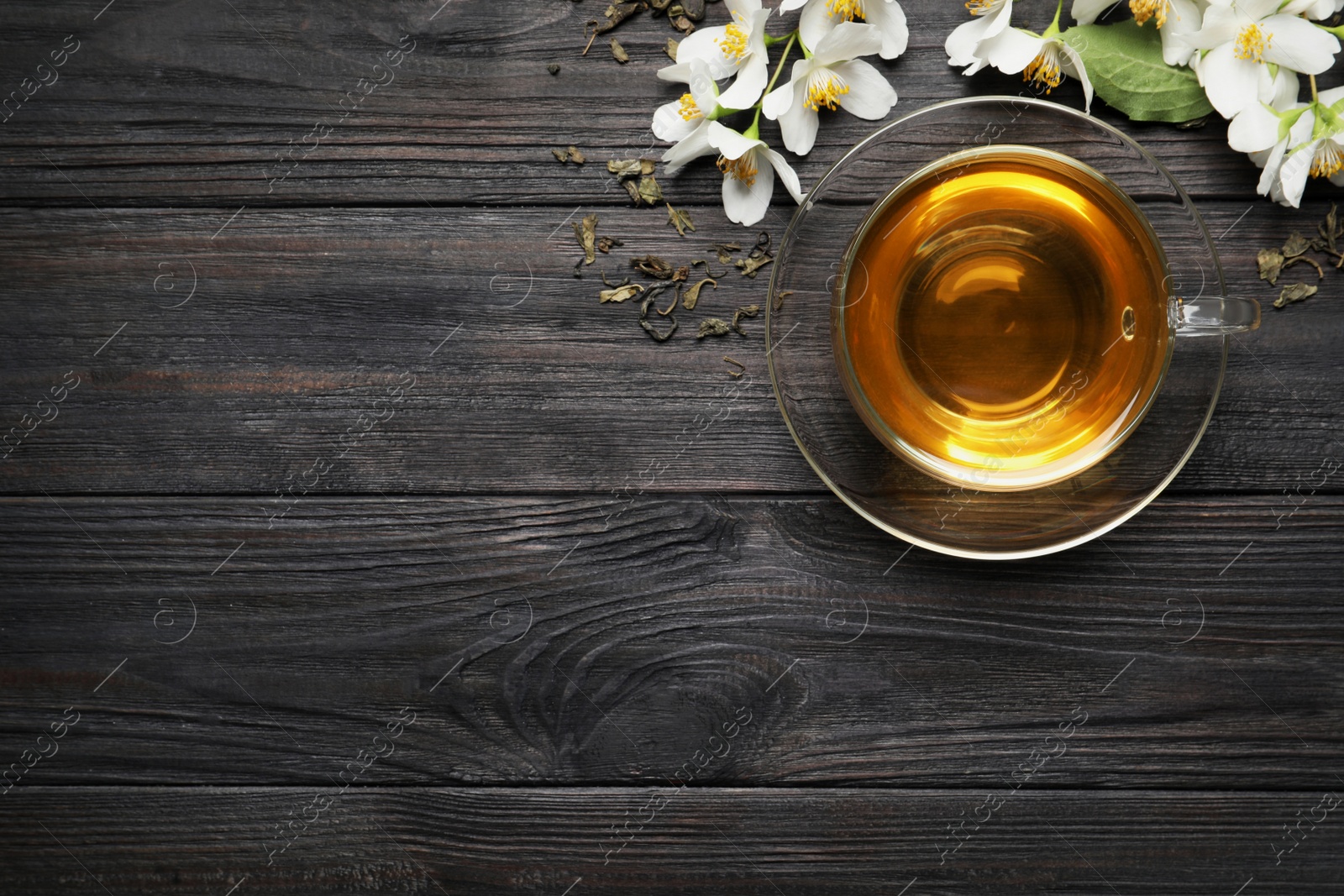 Photo of Glass cup of aromatic jasmine tea, dry leaves and fresh flowers on black wooden table, flat lay. Space for text