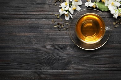 Glass cup of aromatic jasmine tea, dry leaves and fresh flowers on black wooden table, flat lay. Space for text