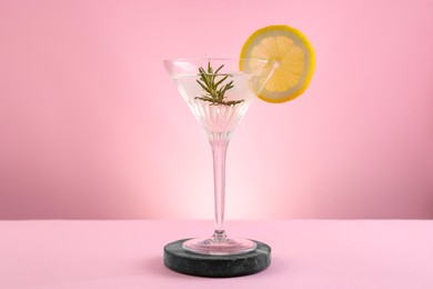 Martini glass of cocktail with lemon slice and rosemary on stand against pink background