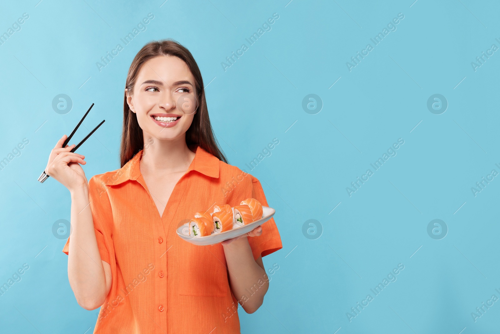 Photo of Happy young woman with plate of sushi rolls and chopsticks on light blue background. Space for text