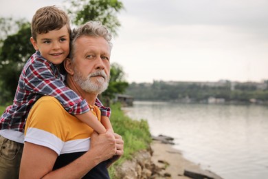 Cute little boy and grandfather spending time together near river, space for text