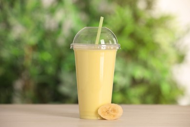 Photo of Plastic cup with tasty smoothie and slice of fresh banana on wooden table outdoors