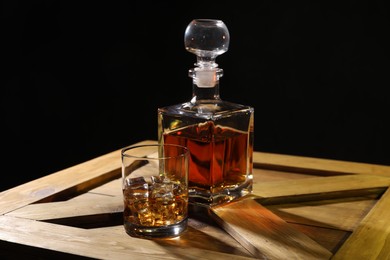 Whiskey with ice cubes in glass and bottle on wooden crate against black background