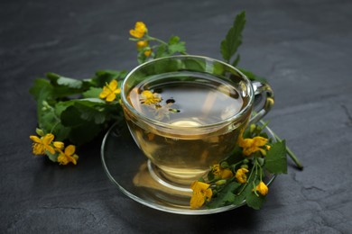 Photo of Glass cup of aromatic celandine tea and flowers on black table