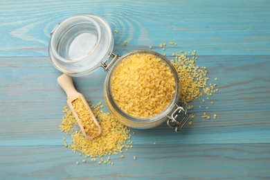 Photo of Glass jar and scoop with raw bulgur on light blue wooden table, flat lay