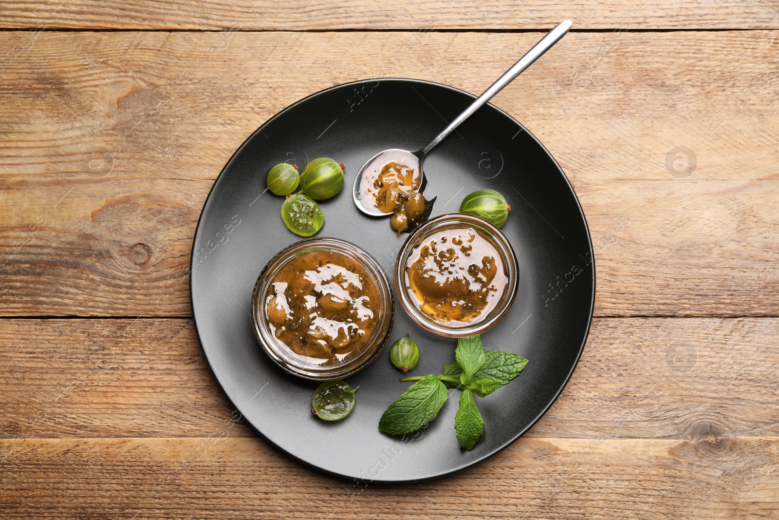 Photo of Plate with jars of delicious gooseberry jam, fresh berries and mint on wooden table, top view