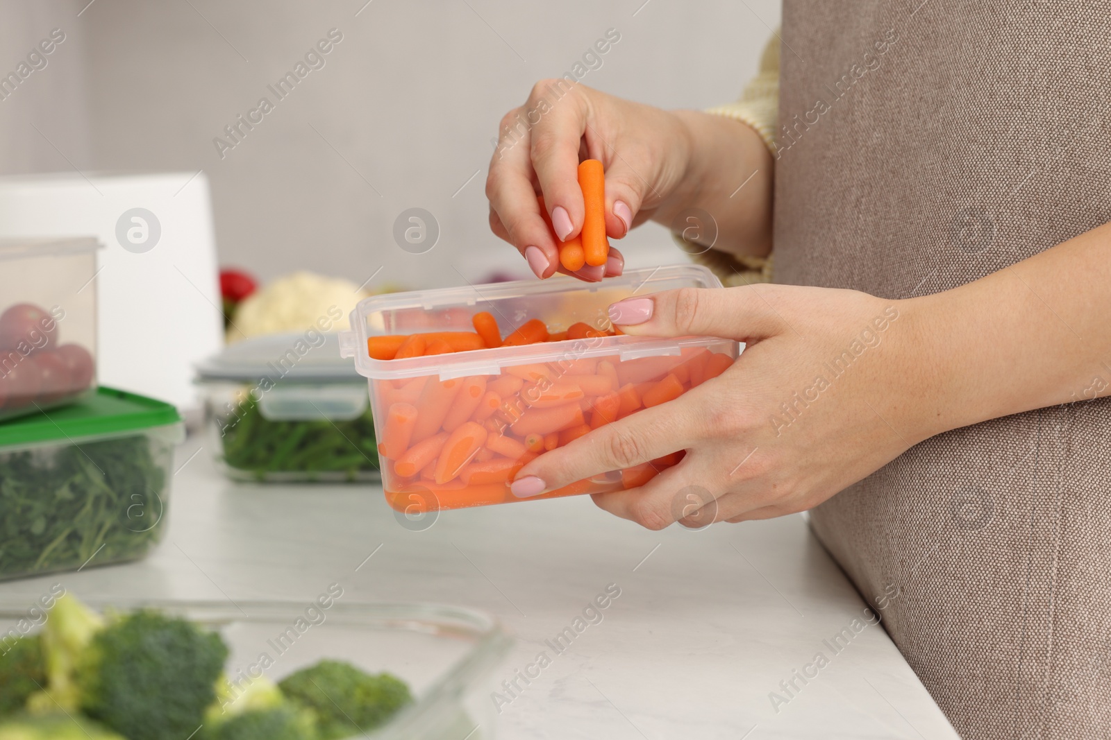 Photo of Woman putting carrots into container with fresh vegetables at white marble table in kitchen, closeup. Food storage