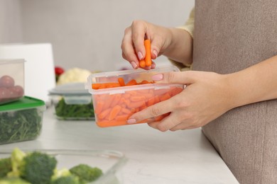 Photo of Woman putting carrots into container with fresh vegetables at white marble table in kitchen, closeup. Food storage