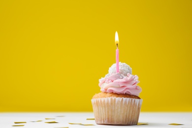 Delicious birthday cupcake with candle and confetti on white table against yellow background. Space for text