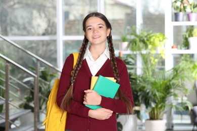 Photo of Teenage girl in school uniform with books and backpack indoors
