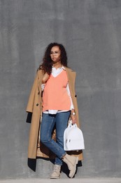 Full length portrait of beautiful African-American woman with stylish white backpack near grey wall outdoors