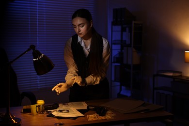 Professional detective wearing protective gloves at table in office at night