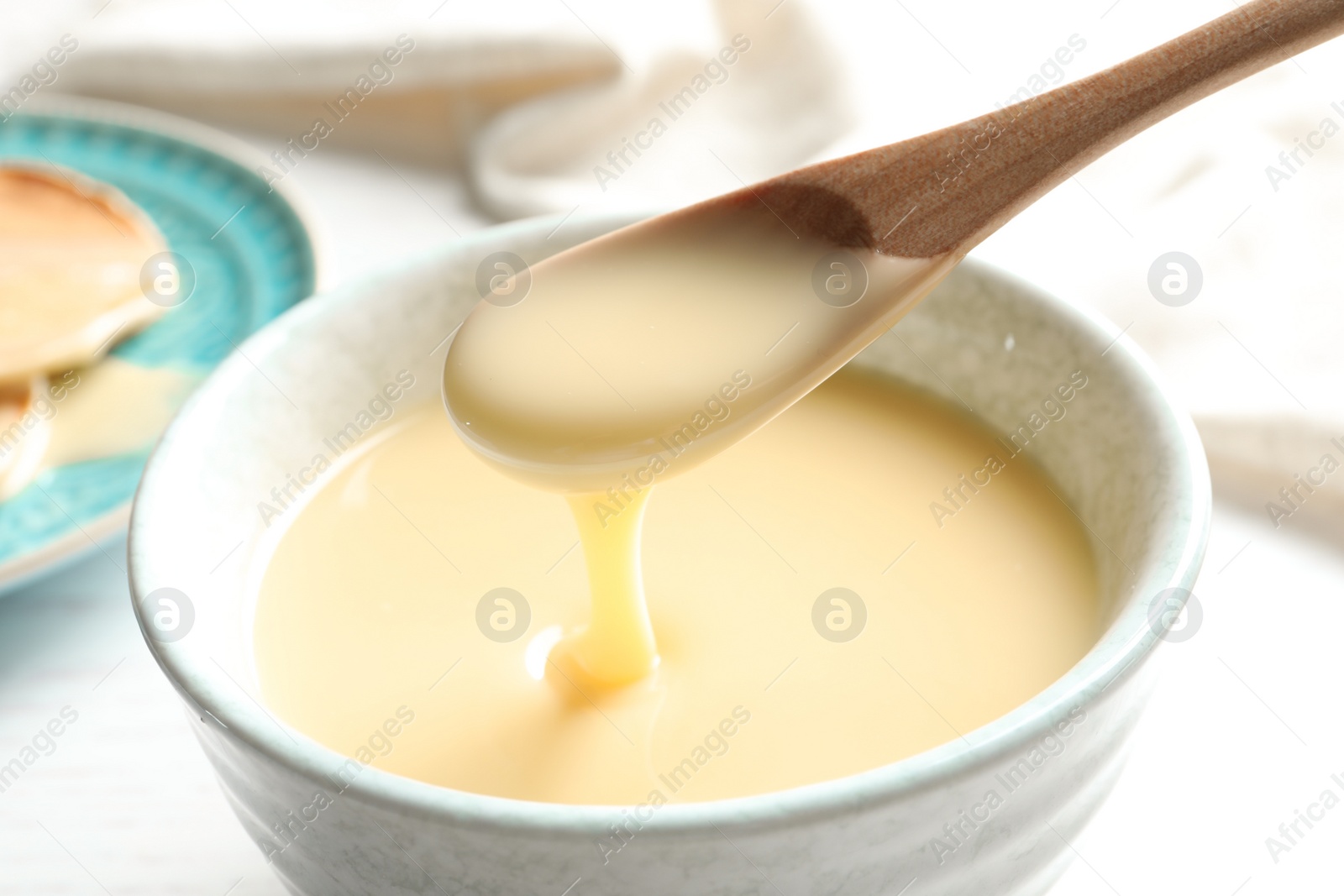 Photo of Spoon of pouring condensed milk over bowl on table, closeup. Dairy products