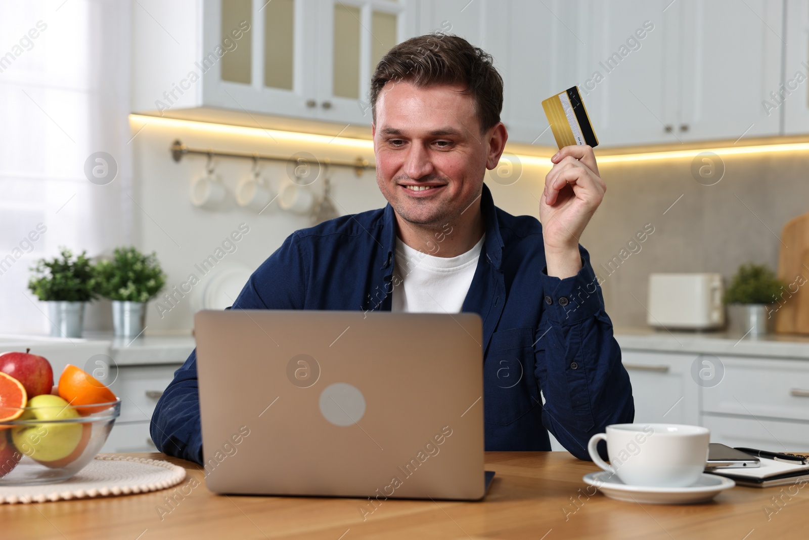 Photo of Happy man with credit card and laptop shopping online at wooden table in kitchen