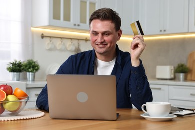 Happy man with credit card and laptop shopping online at wooden table in kitchen