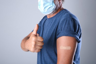 Photo of Senior man in protective mask showing arm with bandage after vaccination on grey background, closeup