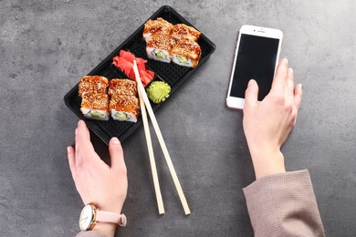 Photo of Woman with smartphone and tasty sushi rolls at grey table, space for text. Food delivery