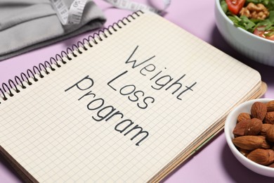 Photo of Notebook with phrase Weight Loss Program, bowl of salad and almonds on pink background, closeup