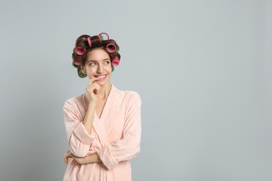 Beautiful young woman in bathrobe with hair curlers on light grey background. Space for text