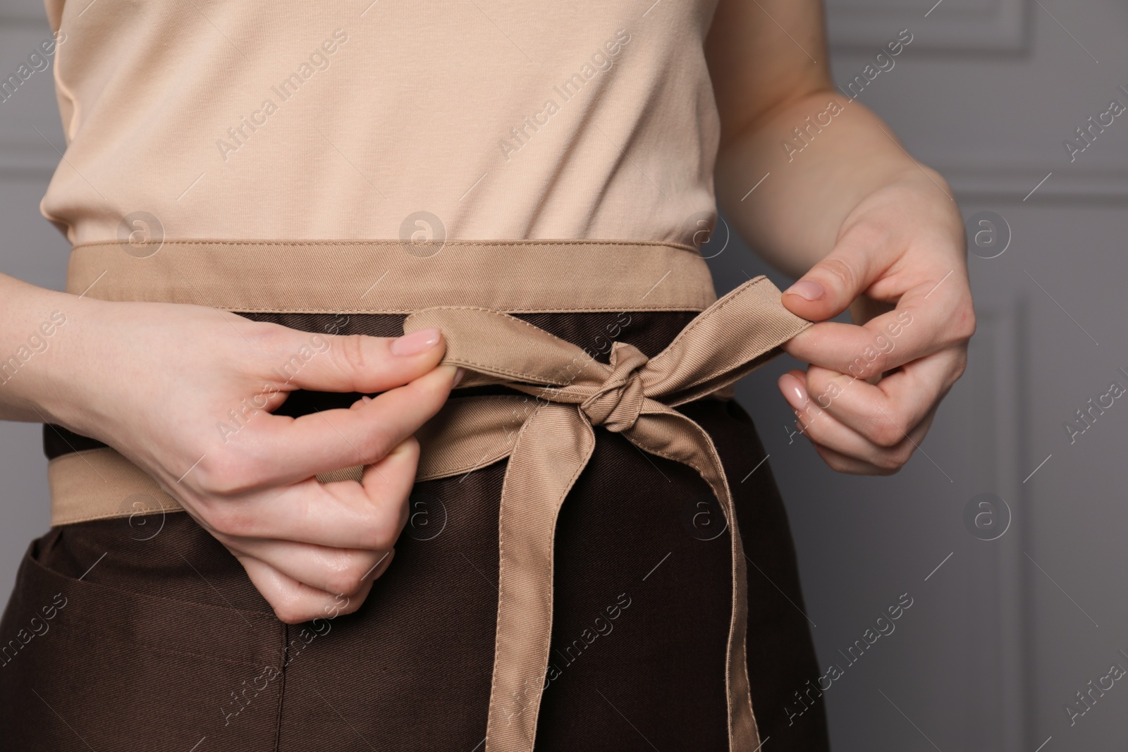 Photo of Woman putting on brown apron against grey wall, closeup