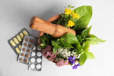 Wooden mortar with fresh herbs, flowers and pills on white table, flat lay