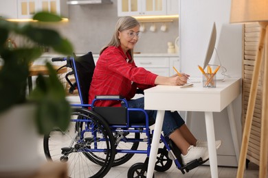 Photo of Woman in wheelchair writing in notebook while using computer at home