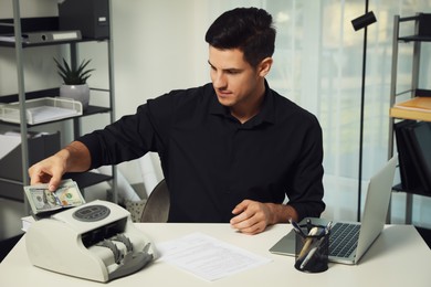 Photo of Man putting money into banknote counter at white table indoors