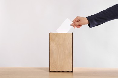 Photo of Woman putting her vote into ballot box on wooden table against light grey background, closeup