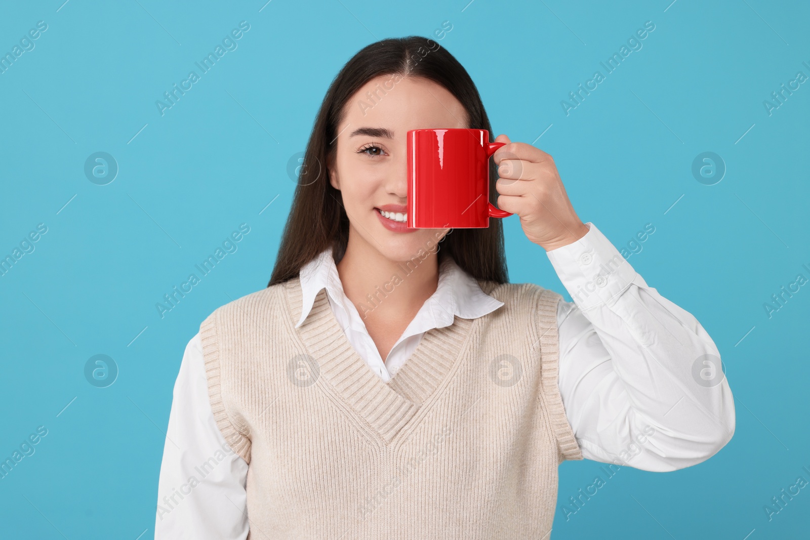 Photo of Happy young woman covering eye with red ceramic mug on light blue background