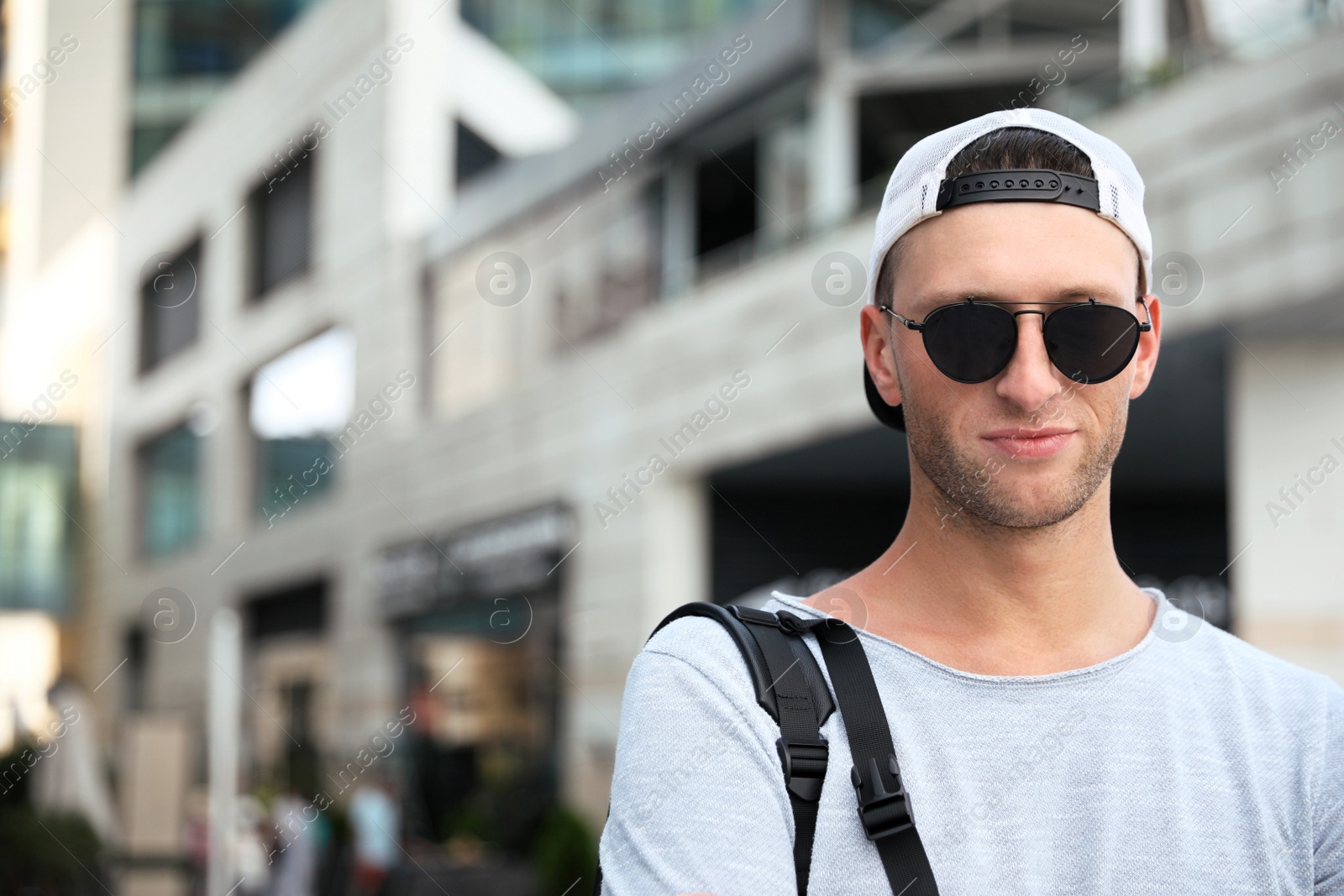 Photo of Handsome young man with stylish sunglasses and backpack near building outdoors, space for text