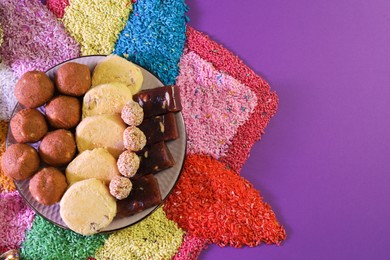 Photo of Diwali celebration. Tasty Indian sweets and colorful rangoli on violet background, flat lay. Space for text