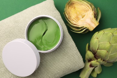 Photo of Package of under eye patches, towel and artichokes on green background, flat lay. Cosmetic product