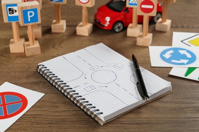Photo of Many different road signs, notebook with sketch of roundabout and toy car on wooden table. Driving school