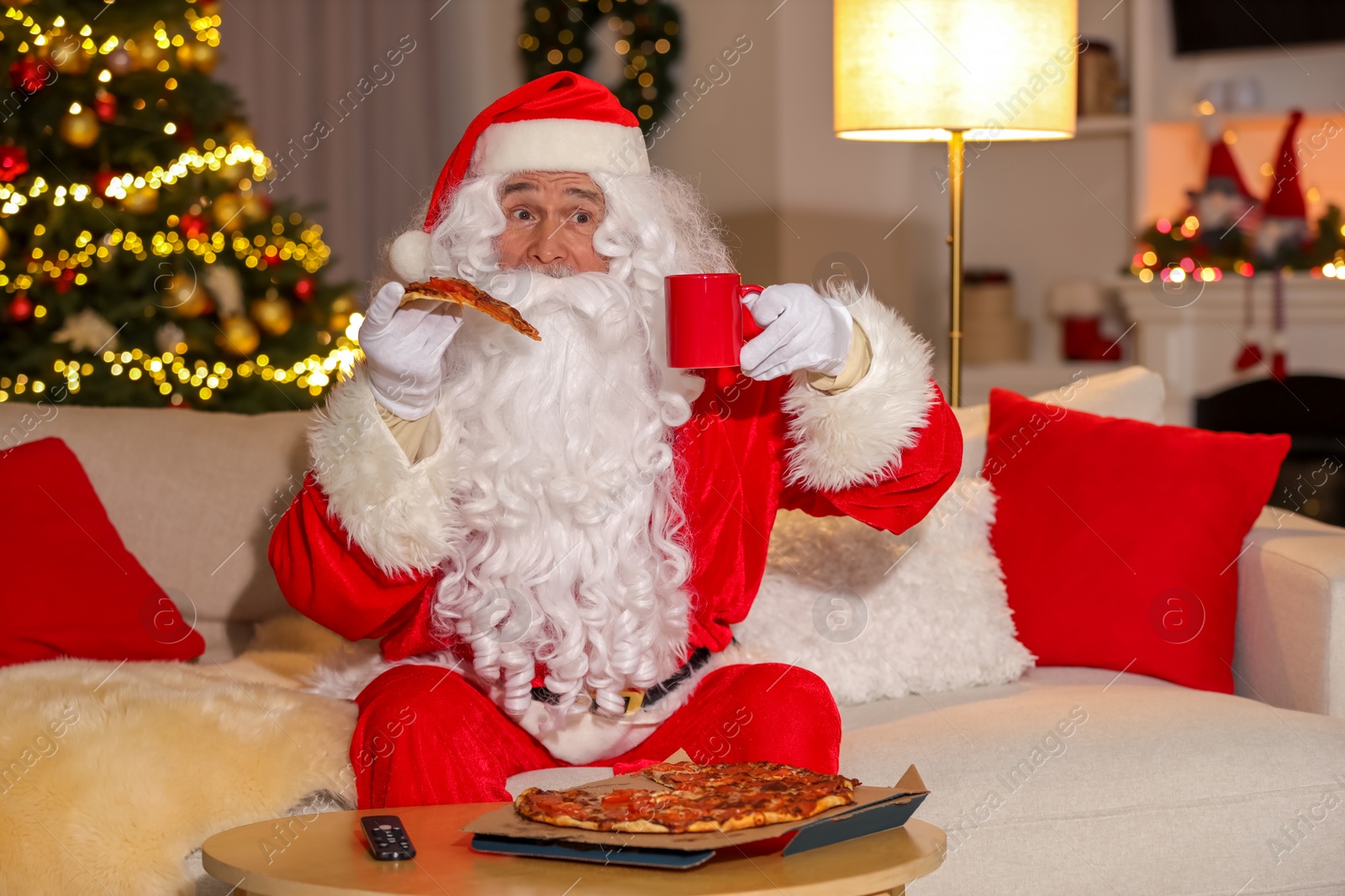 Photo of Merry Christmas. Santa Claus with cup of drink and pizza watching TV on sofa at home