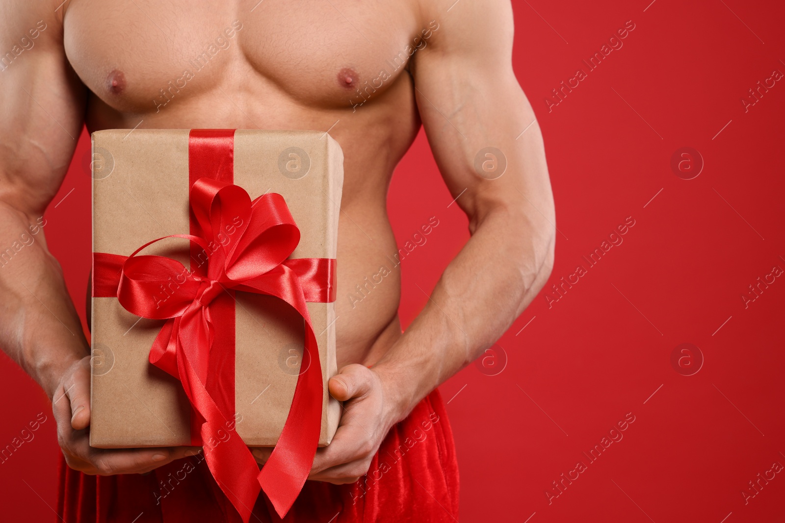 Photo of Young man with muscular body holding Christmas gift box on red background, closeup. Space for text