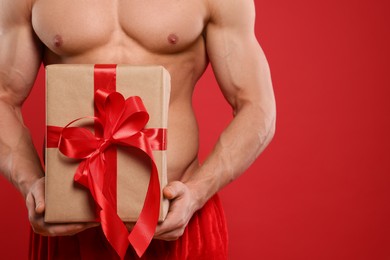 Young man with muscular body holding Christmas gift box on red background, closeup. Space for text