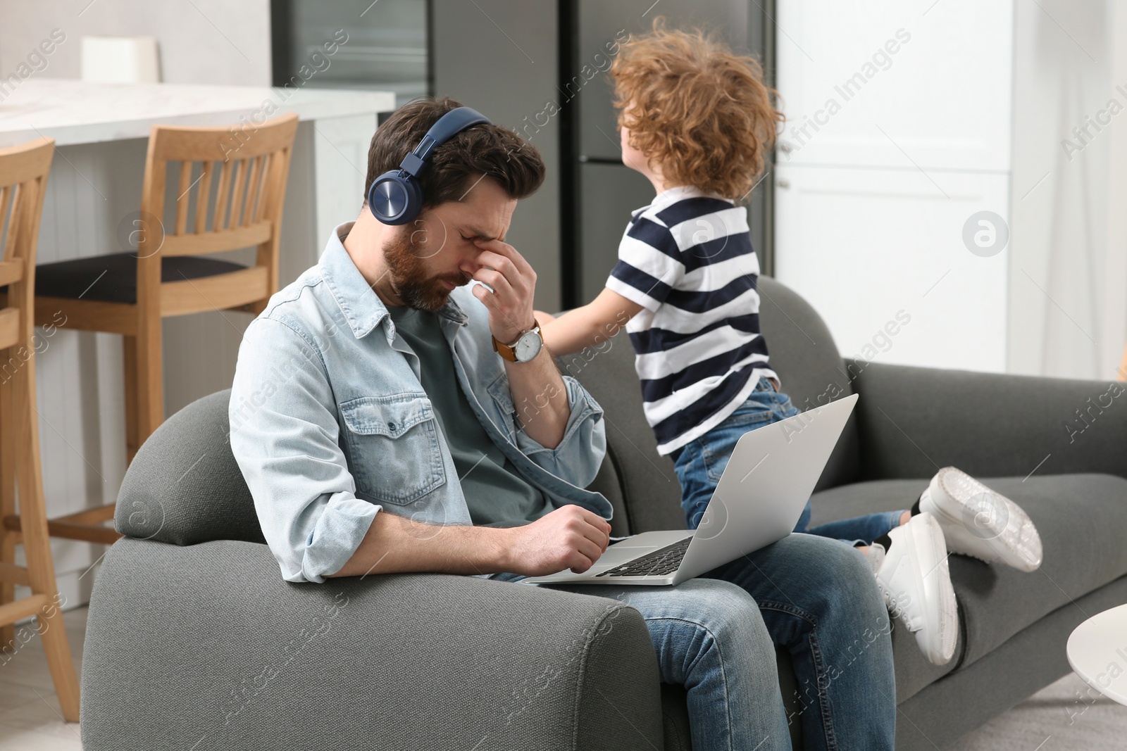 Photo of Little boy bothering his father at home. Man with laptop and headphones working remotely