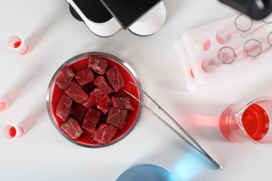 Photo of Petri dish with pieces of raw cultured meat, tweezers and samples on white table, flat lay