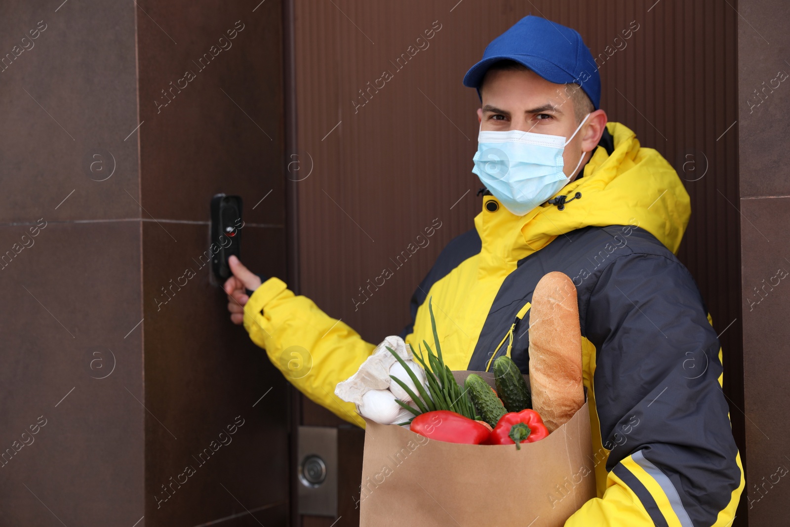 Photo of Courier in medical mask holding paper bag with groceries and ringing doorbell outdoors. Delivery service during quarantine due to Covid-19 outbreak