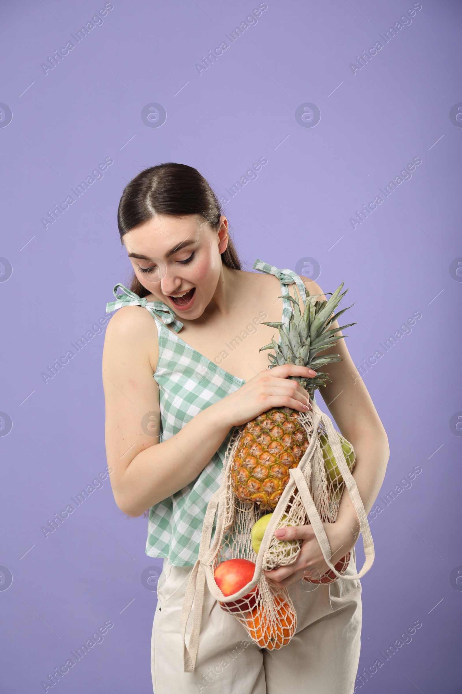 Photo of Surprised woman with string bag of fresh fruits on violet background