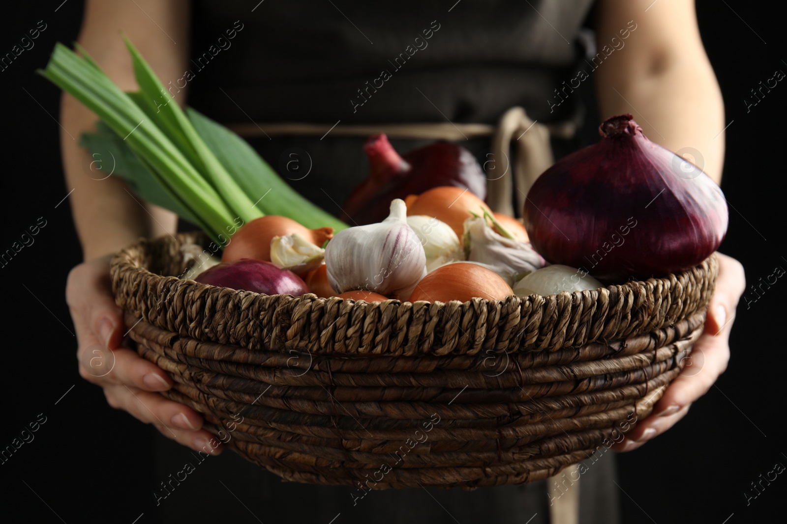 Photo of Woman holding basket with fresh onion bulbs, leeks and garlic on black background, closeup