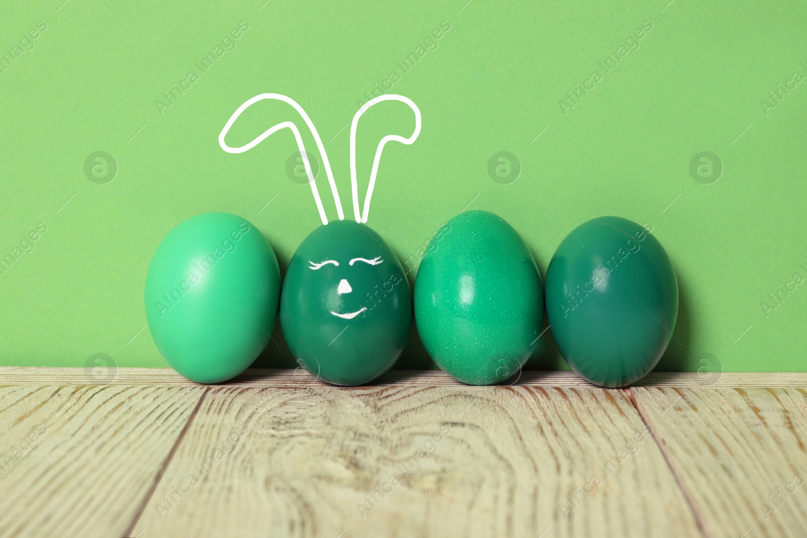 Image of One egg with drawn face and ears as Easter bunny among others on white wooden table against green background