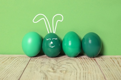 One egg with drawn face and ears as Easter bunny among others on white wooden table against green background