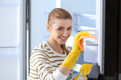 Photo of Woman in rubber gloves cleaning empty refrigerator with rag at home