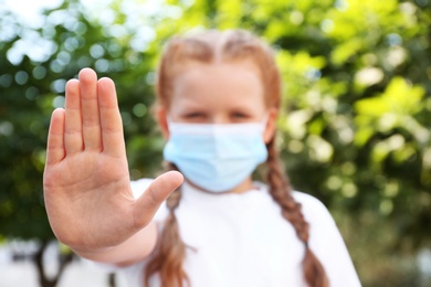 Photo of Little girl in protective mask showing stop gesture outdoors, focus on hand. Prevent spreading of coronavirus