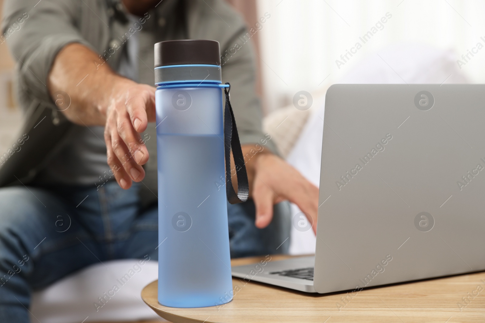 Photo of Man taking transparent plastic bottle of water while working on laptop at home, closeup